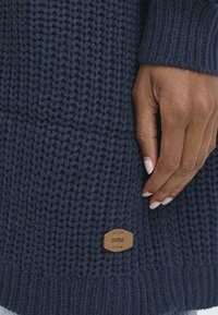 Marineblå strikket sweater med et tekstureret design, ribbede manchetter og et tan læderlogo ved sømmen. Blødt stof med en kraftig vævning.