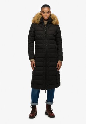 Lange zwarte pufferjas met een bont-gevoerde capuchon, gequilt ontwerp, ritsluiting, zijzakken en geribbelde manchetten, gedragen met blauwe jeans en laarzen.