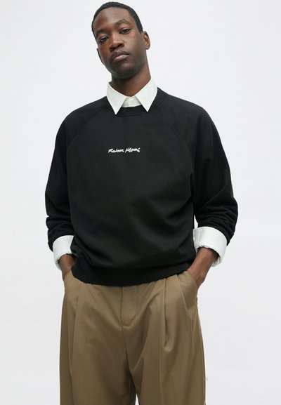 Bărbat purtând un hanorac negru cu textul „Maison Kitsuné”, cămașă albă cu guler și pantaloni bej, cu mâinile în buzunare, fundal neutru.