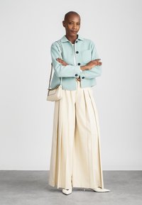 Krátký rukáv mátově zelený límečkový kabát s předními kapsami, smetanové plisé široké kalhoty a malá krémová kabelka přes rameno.