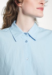Camisa de botão azul clara com colarinho clássico e dois botões visíveis na frente. O tecido parece suave, com uma textura ligeiramente amarrotada.
