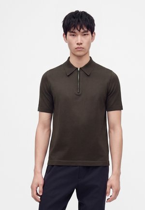 Giovane uomo che indossa una polo a maniche corte marrone scuro con cerniera frontale e pantaloni scuri, in piedi davanti a uno sfondo chiaro e uniforme.