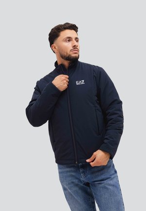 Giacca puffa blu navy realizzata in tessuto morbido, con colletto alto, zip frontale e logo sul petto. Progettata per garantire calore e comfort.
