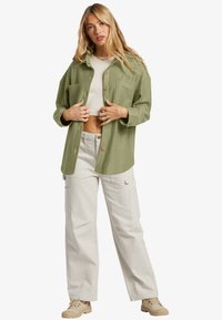 Grön knappjacka i texturerad väv, bärs över en vit crop top och beige cargobyxor, kombinerat med ljusbruna skor.