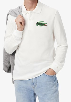 Mężczyzna w białej koszuli polo z długim rękawem i zielonym logo krokodyla, trzyma szary sweter na ramieniu i ma na sobie jasnoniebieskie dżinsy.