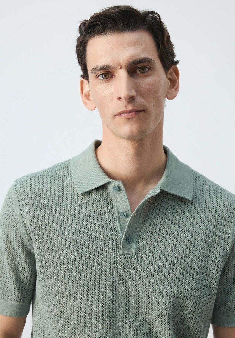 Homme aux cheveux courts et foncés portant un polo en maille vert clair à manches courtes, face à un fond blanc uni.