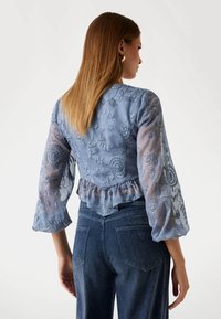 Lichtblauwe geborduurde blouse met doorschijnende, poffende mouwen en gerimpelde zoom; heeft bloemmotieven en een getailleerde achterkant. Gepaard met donkere denim jeans.