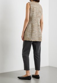 Lätt texturerad, ärmlös beige väst med fransade kanter, kombinerad med mörkgrå, avsmalnande byxor och platta svarta skor, sedd bakifrån.