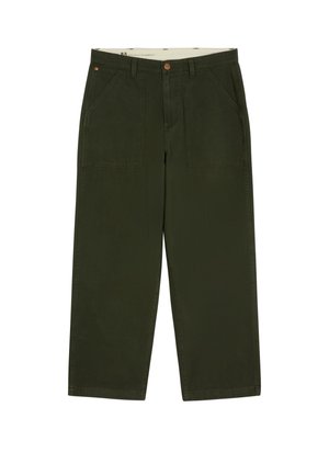 Jeune homme portant une veste boutonnée vert foncé et un pantalon assorti avec des chaussures marron, debout les mains dans les poches sur un fond blanc.