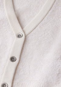 Cardigan color crema con un corpo texturizzato e peloso, scollo a V a costine e cinque bottoni metallici lungo il davanti per la chiusura.