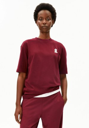 Vrouw met kort krullend haar die een bordeauxrode korte mouwen shirt en bijpassende broek draagt, staand met één hand in de zak tegen een witte achtergrond.