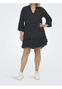 Schwarzes Kleid mit Punkten, V-Ausschnitt und drei Viertel-Ärmeln. Verfügt über einen gestuften Rüschensaum. Kombiniert mit weißen Sneakern.