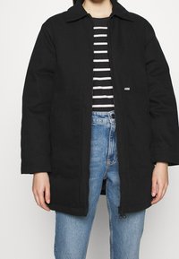 Sort jakke med krave og lynlåslukning foran, med to sidelommer og et diskret logo-mærkat, kombineret med en sort-hvidstribet skjorte og blå jeans.