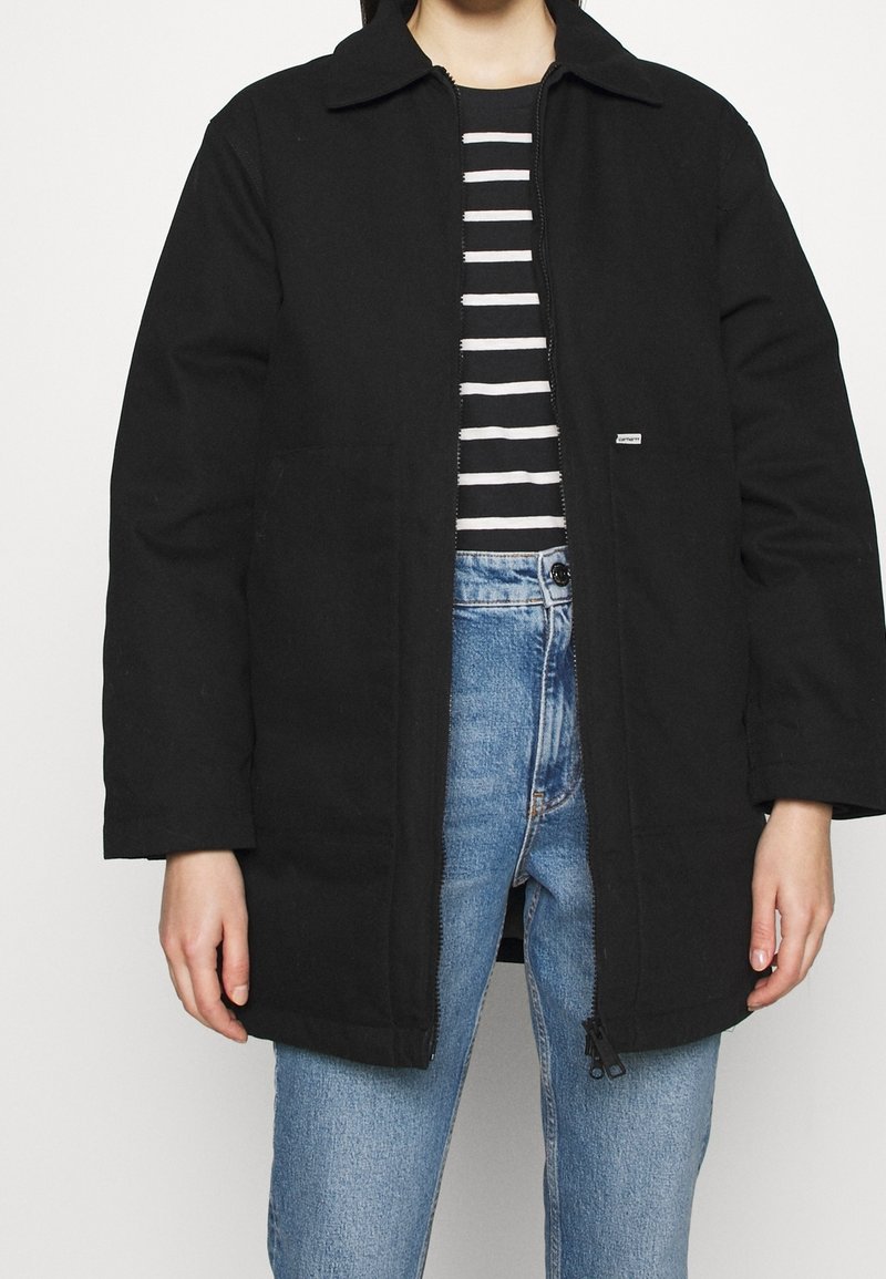 Sort jakke med krave og lynlåslukning foran, med to sidelommer og et diskret logo-mærkat, kombineret med en sort-hvidstribet skjorte og blå jeans.