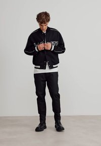 Svart varsityjacka med vitt logo, ribbade muddar och fåll. Kombinerad med en vit skjorta och svarta byxor, avslutad med svarta stövlar.