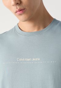 Šviesiai mėlynas medvilninis marškinėlis su apvaliu kaklu ir baltu spaudiniu, kuriame parašyta "Calvin Klein Jeans Įkurtas 1978 metais."