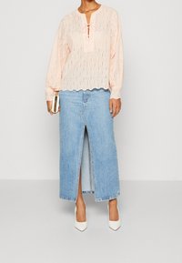 Femme portant un chemisier en dentelle rose clair à manches longues, une jupe en jean bleu clair taille haute avec une fente à l'avant, des talons blancs et tenant une pochette dorée.