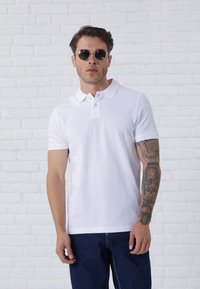 Hvid polo shirt med krave og tre knapper, parret med blå denim jeans. Model bærer sorte runde solbriller og har en detaljeret tatovering på armen.