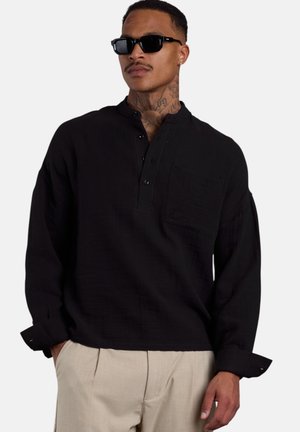 Mann mit Hals-Tattoos trägt ein schwarzes, knöpfbares langärmliges Hemd, beige Hose und schwarze Sonnenbrille, steht mit den Händen in den Taschen.