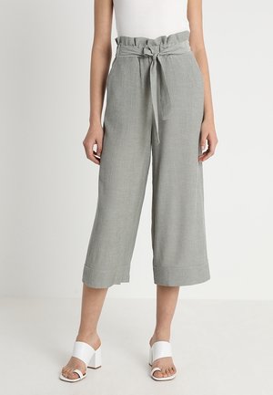 Femme portant un pantalon gris taille haute large et court avec une ceinture nouée, associé à des sandales blanches à talons blocs ouverts à l'avant.