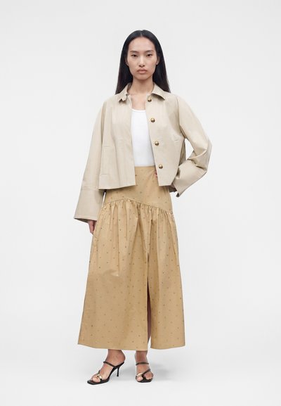 Γυναίκα με μακριά μαλλιά φοράει μπεζ cropped σακάκι με χρυσά κουμπιά, λευκό αμάνικο τοπ, καφέ φαρδιά φούστα με μικρά σχέδια και μαύρες γόβες.