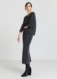 Pull en maille grise à manches longues et coupe décontractée, associé à une jupe longue grise assortie avec une fente latérale. Bottes noires à talons hauts.