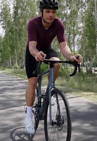 Radfahrer in einem kastanienbraunen Trikot und schwarzen Shorts fährt ein graues Rennrad mit Scheibenbremsen auf einem von Bäumen gesäumten Weg.