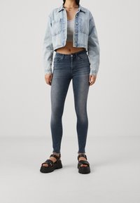 Ljusblå denimjacka i cropped-stil över en grå topp, matchad med mörka skinny jeans och svarta kraftiga sandaler. Har fickor och slitna detaljer.