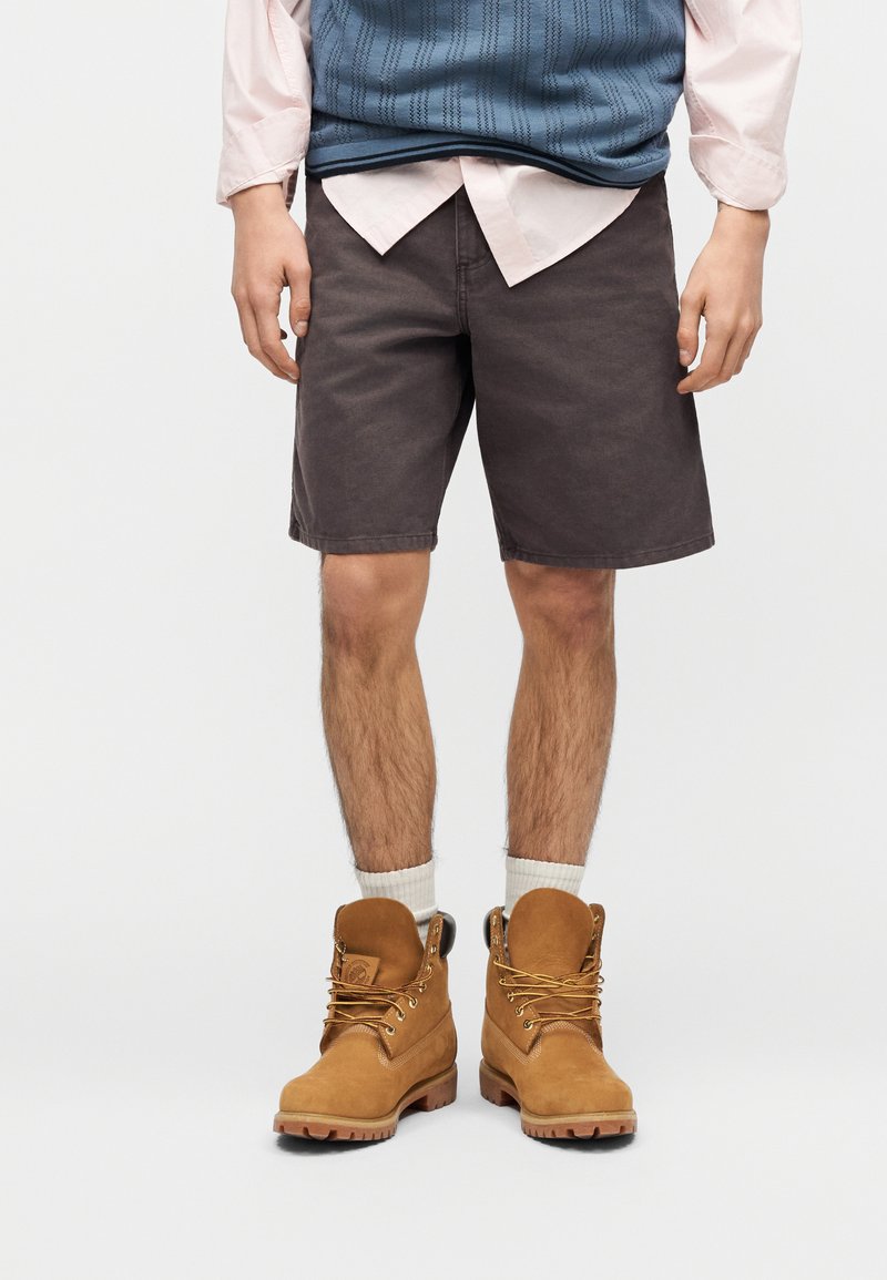 Man draagt donkerbruine korte broek, witte sokken, bruine werkschoenen, een lichtroze overhemd en een blauwe mouwloze trui met een gestructureerd patroon.