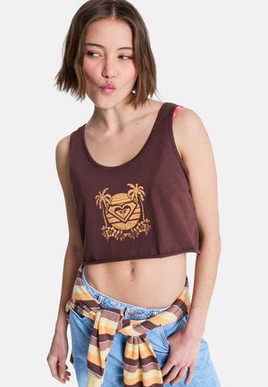 Jeune femme aux cheveux courts bruns portant un crop top marron avec un motif jaune de palmier et de cœur, un jean en denim et un pull rayé noué autour de la taille.