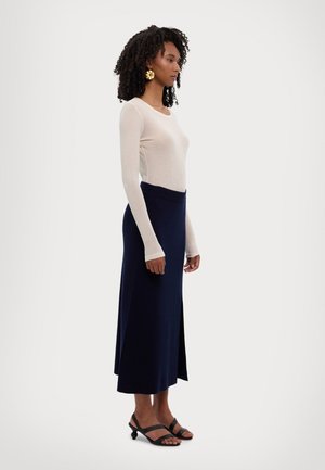 Bluză crem cu mâneci lungi și fustă maxi bleumarin cu slit lateral; modelul poartă sandale negre cu un design distinctiv al tocului.
