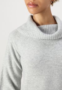 Suéter cinza claro com gola alta e textura canelada; apresenta mangas longas e um acabamento de tecido suave e macio.