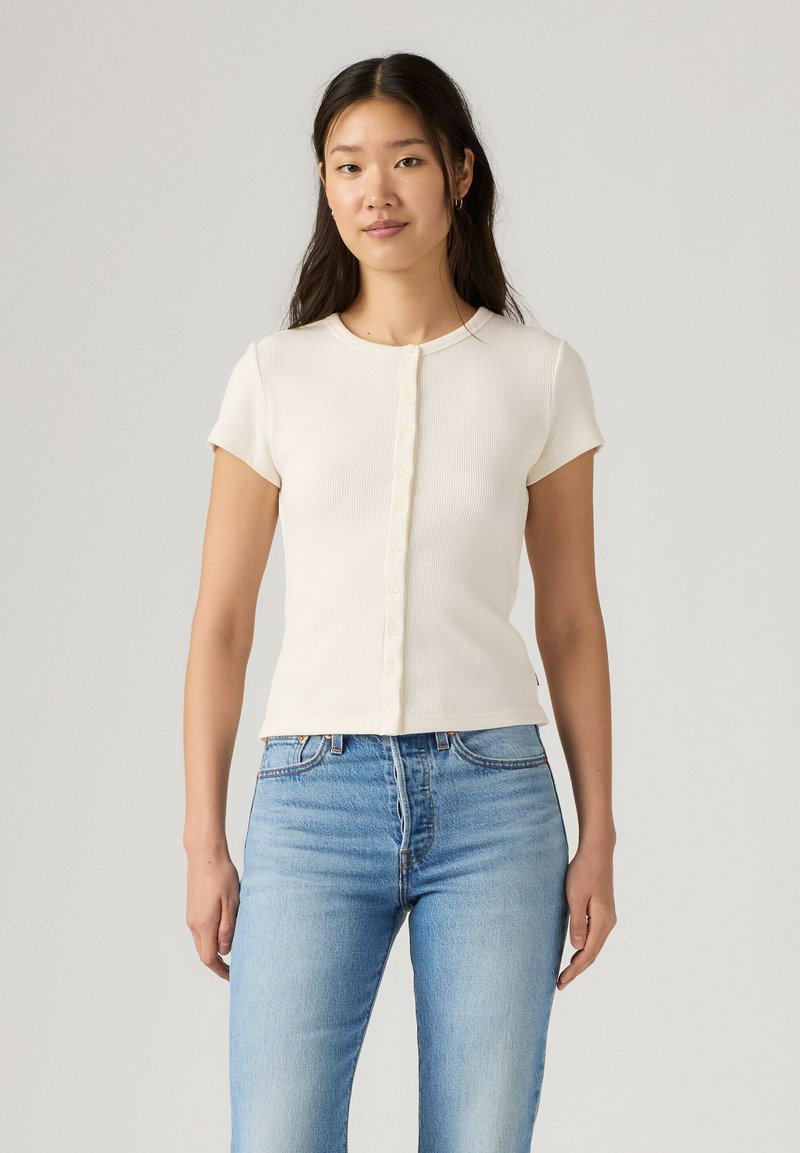 Jeune femme aux longs cheveux foncés portant un haut blanc à manches courtes boutonné et un jean bleu clair, debout devant un fond clair et uni.