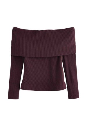 Femme portant un top marron épaules dénudées, pantalon plissé noir taille haute avec une ceinture et des chaussures bordeaux pointues sur fond rouge.