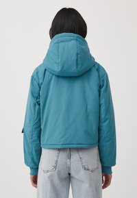 Turkusowa kurtka puchowa z dużym kapturem, krótki fason, ściągacze przy mankietach i gładka, błyszcząca faktura. Noszona z jasnoniebieskimi, wysokimi jeansami.