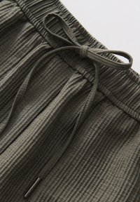 Taille élastique avec cordon noué sur pantalon vert en tissu côtelé montrant un matériau texturé et des extrémités de cordon en métal.