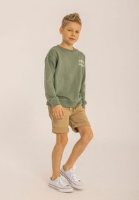 Felpa verde con testo bianco, pantaloni corti beige e sneakers bianchi. Materiale in cotone liscio, vestibilità rilassata, design casual e maniche arrotolate.