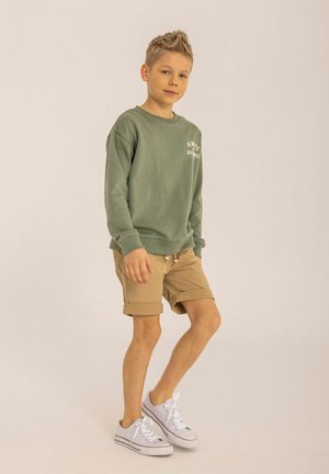 Felpa verde con testo bianco, pantaloni corti beige e sneakers bianchi. Materiale in cotone liscio, vestibilità rilassata, design casual e maniche arrotolate.