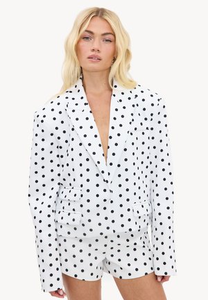 Femme blonde portant un blazer blanc oversize et un short assorti à pois noirs, debout devant un fond uni.