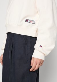Biała, krótka bluza z miękkiego materiału, z naszywką logo w kolorze czerwonym i niebieskim, z prążkowanymi mankietami i dołem, w połączeniu z ciemnymi spodniami.