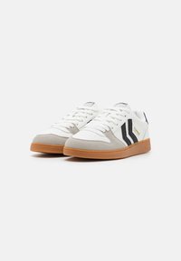 White sneakers featuring a leather upper with grey suede accents, black stripes, rubber gum sole, and lace closure.
