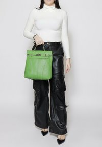 Groene leren handtas met een gestructureerde vorm, bovenhandvat en zilverkleurige hardware. Model draagt een witte coltrui en zwarte leren cargobroek.