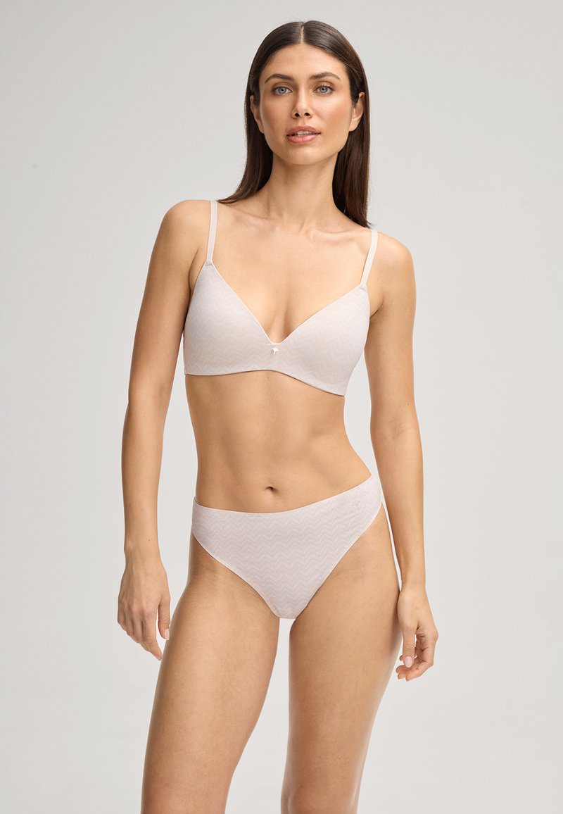 Femme portant un soutien-gorge à motifs beige clair et un sous-vêtement taille haute assorti, se tenant contre un fond neutre uni, face à l'avant.