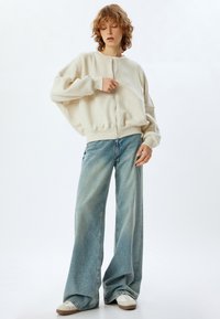 Persona con cabello rizado que lleva un suéter beige de gran tamaño con cremallera, pantalones anchos de mezclilla azul y zapatillas blancas sobre un fondo blanco.