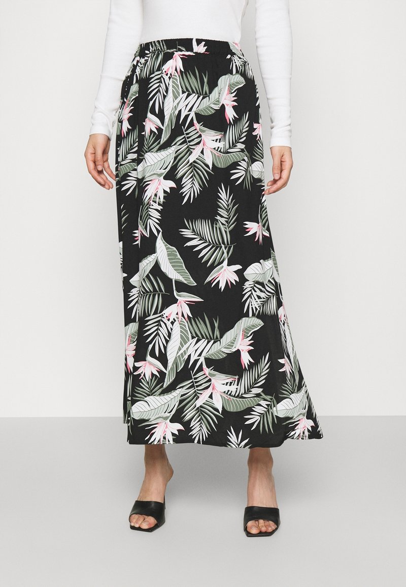 Femme portant une jupe longue noire avec des motifs floraux roses et blancs ainsi que des feuilles vertes, associée à des talons ouverts noirs et un haut blanc à manches longues.