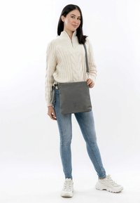 Gray crossbody bag with a smooth texture, zippered closure, and decorative tassels. Simple, minimalist design with a logo accent.