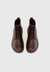 Botas de cuero marrón hasta el tobillo con un acabado texturizado, punteras redondas, cordones y una suela de goma gruesa. La vista superior muestra los ojales y detalles de costura.