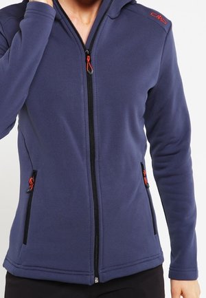 Person iført en mørkeblå lynlåsjakke med orange lynlåsdragere og en rød logo på skulderen, stående foran en ensfarvet baggrund.