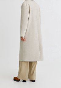 Abrigo de lana beige con cuello, mangas largas y silueta recta. Llevado sobre pantalones de pierna ancha beige a rayas y zapatos de plataforma marrones.
