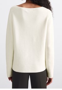 Femme aux cheveux bouclés portant un pull à manches longues écru et un pantalon foncé, vue de dos sur un fond uni.